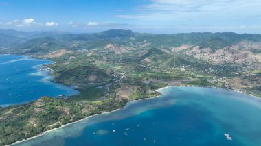 Yeşil tepelerin arasına yerleşmiş bir sahil köyünün havadan çekilmiş görüntüsü. Uzakta yükselen dağlar ve kıyı boyunca uzanan sakin mavi körfezler..