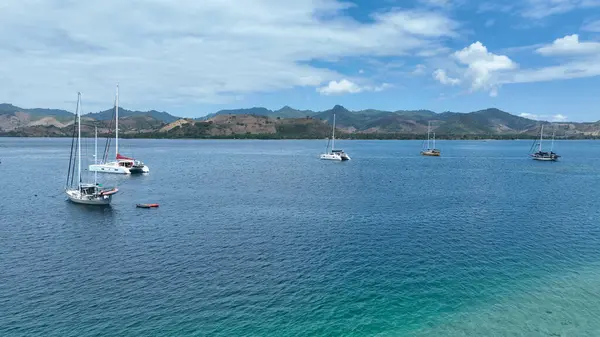 Yelkenliler ve katamaranlar arka planda yuvarlanan yeşil tepelerle derin mavi sularda huzur içinde yüzerler. Sahne, açık denizde sükunet ve macera saçar..