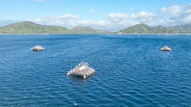 Bir bambu balıkçı salları kümesi Lombok açıklarında turkuaz sularda sürükleniyor. Manzarada dağlar ve parlak bir gökyüzünün altında uzak bir kıyı şeridi var..