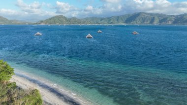 Suda yüzen geleneksel bambu balığı platformlarıyla turkuaz bir körfezin havadan görüntüsü. Arkaplanda el değmemiş kumlu bir sahil ve yemyeşil tepeler.
