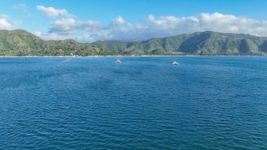 Kristal mavi deniz ufukta iki bambu balıkçılık platformuyla dramatik yeşil dağlar ve dağınık bulutlarla dolu canlı mavi bir gökyüzü ile uzanır..