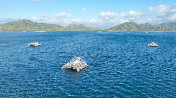 Bir bambu balıkçı salları kümesi Lombok açıklarında turkuaz sularda sürükleniyor. Manzarada dağlar ve parlak bir gökyüzünün altında uzak bir kıyı şeridi var..