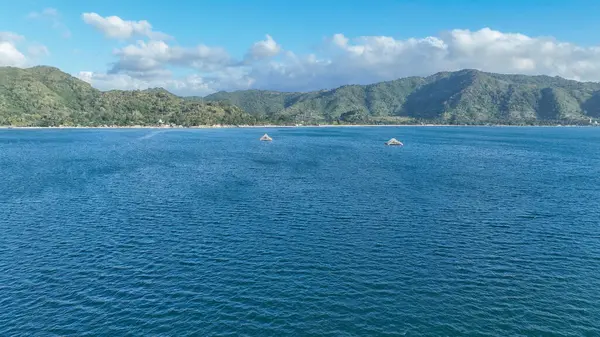 Kristal mavi deniz ufukta iki bambu balıkçılık platformuyla dramatik yeşil dağlar ve dağınık bulutlarla dolu canlı mavi bir gökyüzü ile uzanır..