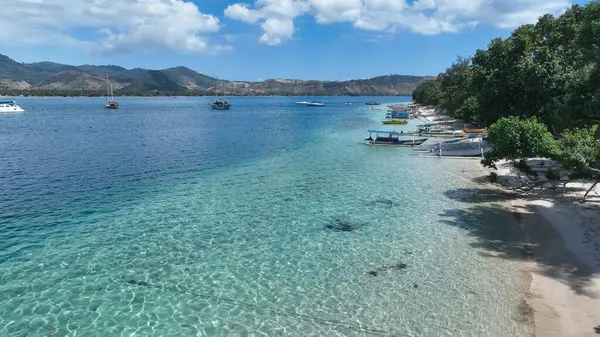 Temiz sığ sular, kumlu deniz tabanını, teknelerle benekli, güneşli gökyüzünün altında yükselen manzaralı dağlarla ortaya çıkarır..