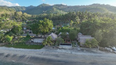 Sazlı çatıları olan geleneksel bungalovlar, arka planda yükselen Lomboks görkemli tepeleriyle, palmiyeler ve yemyeşil ağaçlarla gölgelenmiş sessiz kumlu bir plaj hizasında..