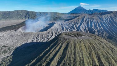 Bromo Dağı ve komşu Batok volkan konisinin çarpıcı kuş bakışı görüntüsü, çarpıcı tepeler ve volkanik dokular canlı bir gökyüzünün altında ufka doğru uzanıyor..