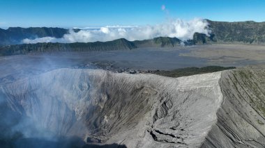Dramatik bir kuş bakışı görüntüsü Bromo Dağı 'nın uçsuz bucaksız kraterini engebeli volkanik manzaranın üzerinde dönen sis ve berrak mavi gökyüzünün altındaki uzak tepeleri gözler önüne serer.