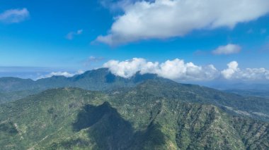 Bulutlar canlı mavi gökyüzünün altındaki tropik bir dağ sırasının verimli zirvelerini nazikçe kucaklıyor. Dramatik gölgeler ve canlı yeşillik macera ve keşfi davet eden nefes kesici bir manzara yaratır..