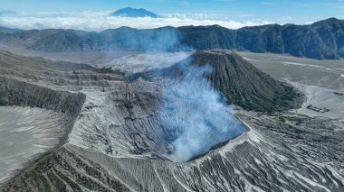 Bromo Dağı 'nın hava manzarası kraterden yükselen dumanı gösteriyor. Dik volkanik yamaçlar ve geniş ovaların üzerindeki uzak dağlar..