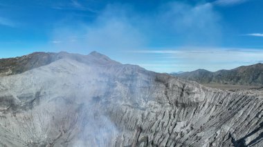 Bir panoramik manzara, uzak dağlara karşı kurulmuş volkanik krateri ve engebeli arazinin üzerinde sürüklenen duman zerreleriyle berrak mavi gökyüzünü yakalar..
