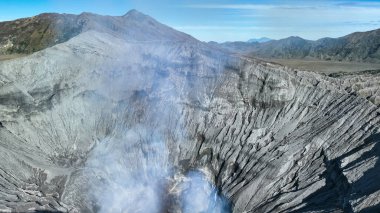 Bromo Dağı 'nın havadan görünüşü geniş Kum Denizi ile çevrili ve yemyeşil dağ sırtlarıyla çevrili volkanik duman bulutu yayan dramatik kraterini gözler önüne seriyor..