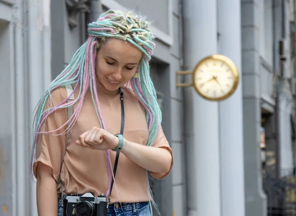Check the time. A young woman looks at her watch while standing against the background of a clock on a city street. Hipster girl with dreadlocks hairstyle with blue hair.
