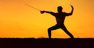 Man is engaged in fencing in an open space against the backdrop of sunset.  Silhouette of a man with a sword.