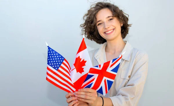微笑的女学生拿着加拿大 英国和美国国旗 在灰墙的背景上看着相机 英语语言教育 — 图库照片