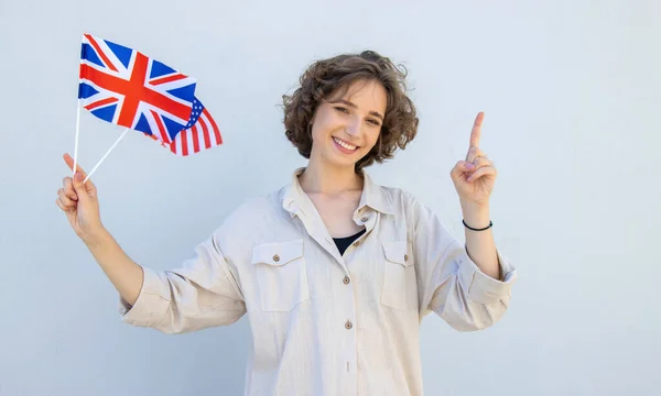 微笑的女学生举着英国和美国国旗 看着相机 用手指指向灰色的墙壁背景 英语语言教育 — 图库照片