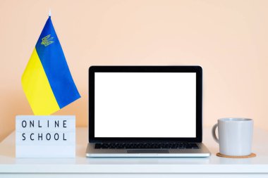 Ukrayna bayrağının yanındaki masada beyaz ekranlı bir laptop ve Online School yazan bir ekran bulunuyor. Uzaktan eğitim. İnternet üzerinden eğitim satışı için sunum düzeni.