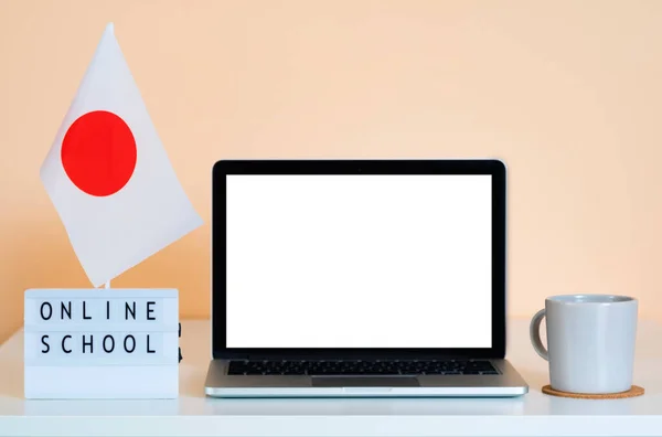 Laptop with blank white screen on table next to Japan flag and display that reads Online School. Distance education. Mockup for selling training via internet or language courses.