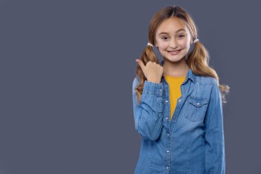 Genç kız parmağını yana doğru uzattı. 10 yaşında, kot gömlekli ve gri arka planda sarı bluzlu bir kız öğrenci..
