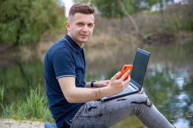 Genç adam temiz havada çalışıyor. Dizüstü bilgisayarı ve akıllı telefonu olan serbest çalışan bir erkek göl kenarında oturuyor..