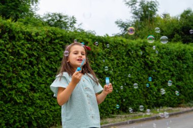 Yeşil bir çitin arkasında sabun köpüğüyle oynayan küçük kız..