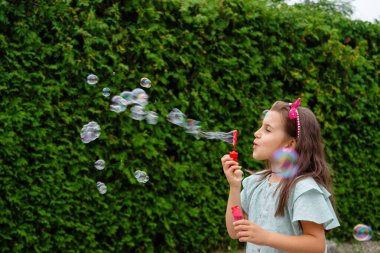Yeşil bir çitin arkasında sabun köpüğüyle oynayan küçük kız..