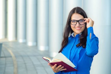 Ofis binasının arka planında, fotokopi alanı olan modern bir iş kadınının portresi. Gülümseyen mutlu öğretmen kadın. Gözlüklü iş kadını, elinde bir günlük ya da kitap tutuyor..