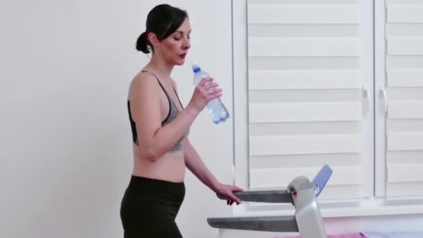 若い女性はトレッドミルで運動し 体を水分補給するために水を飲んでいます ビデオクリップ4K — ストック動画