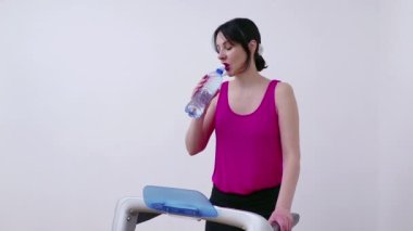 Genç bir kadın koşu bandında egzersiz yapıyor ve vücudunu sulandırmak için su içiyor, Video 4K.