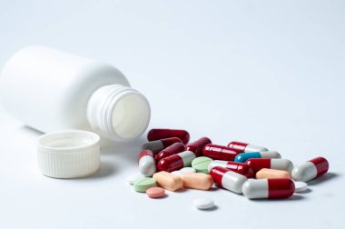 Fotoğraftaki beyaz şişelerin etrafına saçılmış çeşitli hapların, tabletlerin ve kapsüllerin hafif arkaplanlı fotoğrafları.