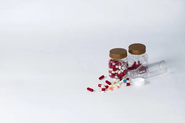 Fotoğraftaki ambalajdaki şeffaf şişelerin etrafına saçılmış çeşitli hapların, tabletlerin ve kapsüllerin hafif arkaplanlı fotoğrafları.