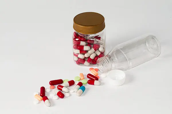 Fotoğraftaki ambalajdaki şeffaf şişelerin etrafına saçılmış çeşitli hapların, tabletlerin ve kapsüllerin hafif arkaplanlı fotoğrafları.