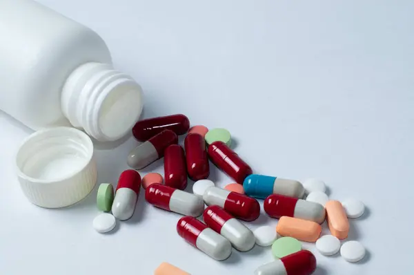 Fotoğraftaki beyaz şişelerin etrafına saçılmış çeşitli hapların, tabletlerin ve kapsüllerin hafif arkaplanlı fotoğrafları.