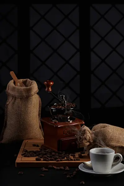 Kahve değirmeninin fotoğrafı, kahve çekirdeği, kahve kaşığı, kahve fincanı ve koyu bir pencere arkasında kahve çekirdeği.