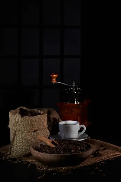Kahve değirmeninin fotoğrafı, kahve çekirdeği, kahve kaşığı, kahve fincanı ve koyu bir pencere arkasında kahve çekirdeği.