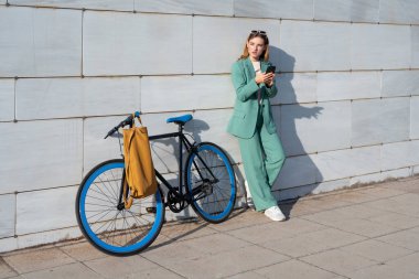 Genç, kızıl saçlı, yeşil takım elbiseli, akıllı telefon kullanan mermer arka planda bisikleti olan bir kadın.