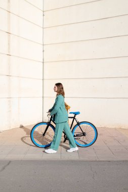 Genç, kızıl saçlı, yeşil takım elbiseli bir kadın. Arka planda modern bir binayla finans bölgesinde bisikletiyle yürüyor.