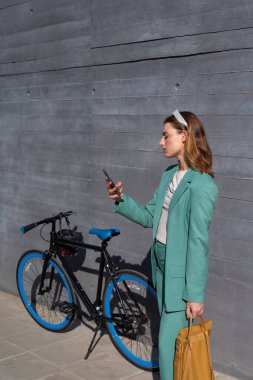 Genç, kızıl saçlı, yeşil takım elbiseli kadın siyah arka planda bisikletiyle akıllı telefon kullanıyor.