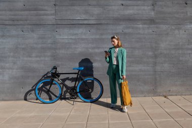 Genç, kızıl saçlı, yeşil takım elbiseli kadın siyah arka planda bisikletiyle akıllı telefon kullanıyor.