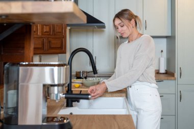 Modern mutfakta ellerini musluk altında yıkayan beyaz bir kadın.