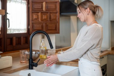 Modern mutfakta ellerini musluk altında yıkayan beyaz bir kadın.