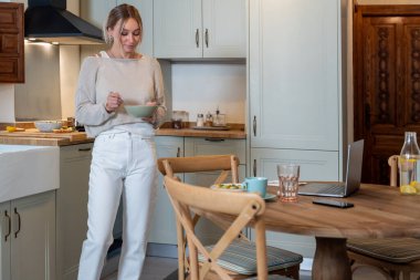 Gündelik kıyafetli beyaz kadın tezgahta duruyor ve modern mutfakta kahvaltıya sağlıklı bir yemek hazırlıyor. 