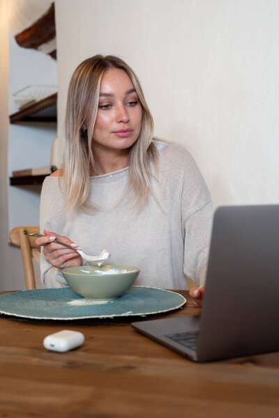 Кавказская женщина ест вкусный завтрак со свежей клубникой, сидя за столом и работая на ноутбуке 