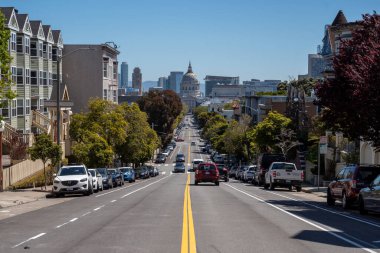 San Francisco, Kaliforniya, ABD - 22 Nisan 2023: Bulutsuz mavi gökyüzüne karşı yeşil ağaçlı, park etmiş arabalı ve asfalt kurbağada araç trafiği olan Belediye Binası ve caddenin tepesinden görüntü