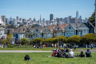 Alamo Square Park, San Francisco, Kaliforniya, ABD - 22 Nisan 2023: Yeşil Park 'ta dinlenen ve bulutsuz mavi gökyüzü altında gün ışığında Boyalı Hanımlar ve Şehre hayran olan mutlu mahalle sakinleri