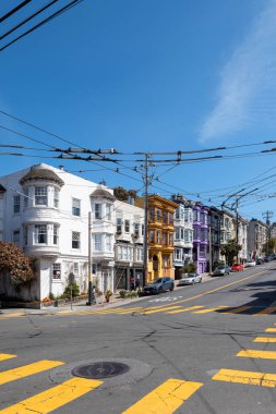 Castro Caddesi, San Francisco, Kaliforniya, ABD - 23 Nisan 2023: Dik sokaktaki renkli evler, asfalt yolda park etmiş arabalar, sarı izler ve bulutlu mavi gökyüzünün altında asılı kablolar