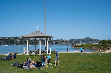 Sausalito, California, ABD - 24 Nisan 2023: Güneşli bir günde deniz suyu yakınlarındaki yeşil çimlerde dinlenen ve eğlenen bir grup insan, binaları ve seyrek bulutlu mavi gökyüzü olan şehirlere karşı.