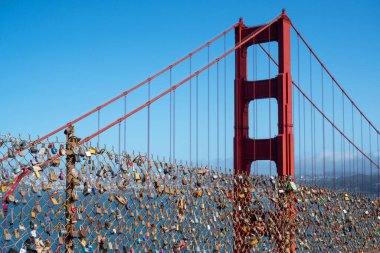 Battery Spencer, San Francisco, Kaliforniya, ABD - 24 Nisan 2023: Metal çitlerin üzerindeki aşk kilitleri ve bulutsuz mavi gökyüzünün altındaki bulanık dağlara karşı gün ışığında Golden Gate Köprüsü süspansiyonu