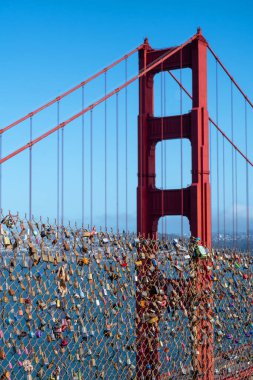Battery Spencer, San Francisco, Kaliforniya, ABD - 24 Nisan 2023: Metal çitlerin üzerindeki aşk kilitleri ve bulutsuz mavi gökyüzünün altındaki bulanık dağlara karşı gün ışığında Golden Gate Köprüsü süspansiyonu