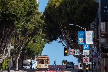 San Francisco, Kaliforniya, ABD - 24 Nisan 2023: Tepe caddesindeki renkli asfalt yolda yeşil ağaçların altından bulutsuz mavi gökyüzüne karşı geçen yol işaretli kablo araba