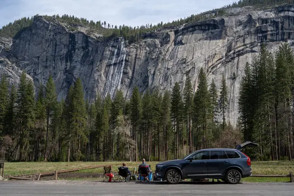 ヨセミテ アメリカ 2023 日当たりの良い木に囲まれた高い雄大な岩山に対して国立公園の車の近くに座っている旅行者 ロイヤリティフリーのストック画像